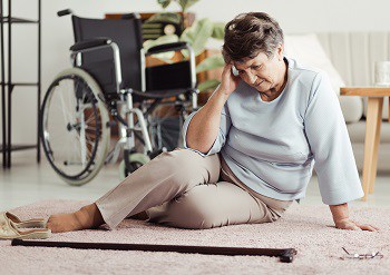 Alte Frau sitzt halbliegend auf einem Teppichboden und hält sich den Kopf, ihre Brille ist zu Boden gefallen.